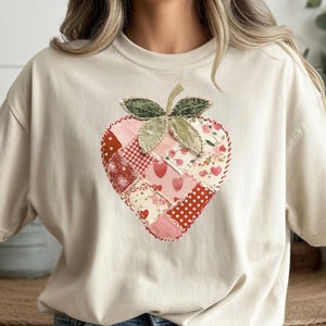 Puede incluir: Camiseta color crema con un diseño de fresa en forma de corazón. La fresa está hecha de tela de retazos en tonos rojos, rosas y blancos, con un tallo y hojas verdes. El diseño está centrado en la camiseta.
