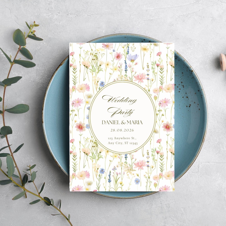 May include: A floral wedding party invitation card with the text "Wedding Party" and the names "Daniel & Maria" and the date "28.08.2026". The card is placed on a blue plate and decorated with a floral pattern.