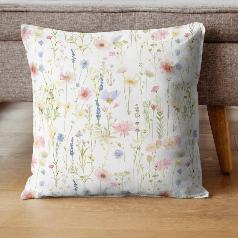 May include: A square throw pillow with a white background, covered in a watercolor floral pattern. The design features a variety of wildflowers in shades of pink, blue, yellow, and purple. The pillow is placed on a wooden floor.