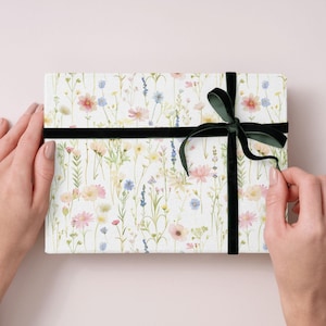 May include: A rectangular gift box wrapped in white paper with a floral pattern of pink, yellow, and blue wildflowers. The box is tied with a black velvet ribbon and bow.