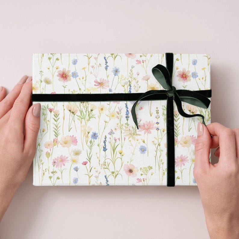 May include: A rectangular gift box wrapped in white paper with a floral pattern of pink, yellow, and blue wildflowers. The box is tied with a black velvet ribbon and bow.