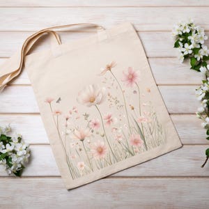 K&ouml;nnte beinhalten: Eine beigefarbene Stofftasche mit einem aufgedruckten Motiv aus zarten Wildblumen in sanften Rosa- und Wei&szlig;t&ouml;nen. Das Design zeigt verschiedene St&auml;ngel, Bl&uuml;tenbl&auml;tter und kleine Bl&auml;tter auf der Vorderseite sowie zwei Tragegriffe.