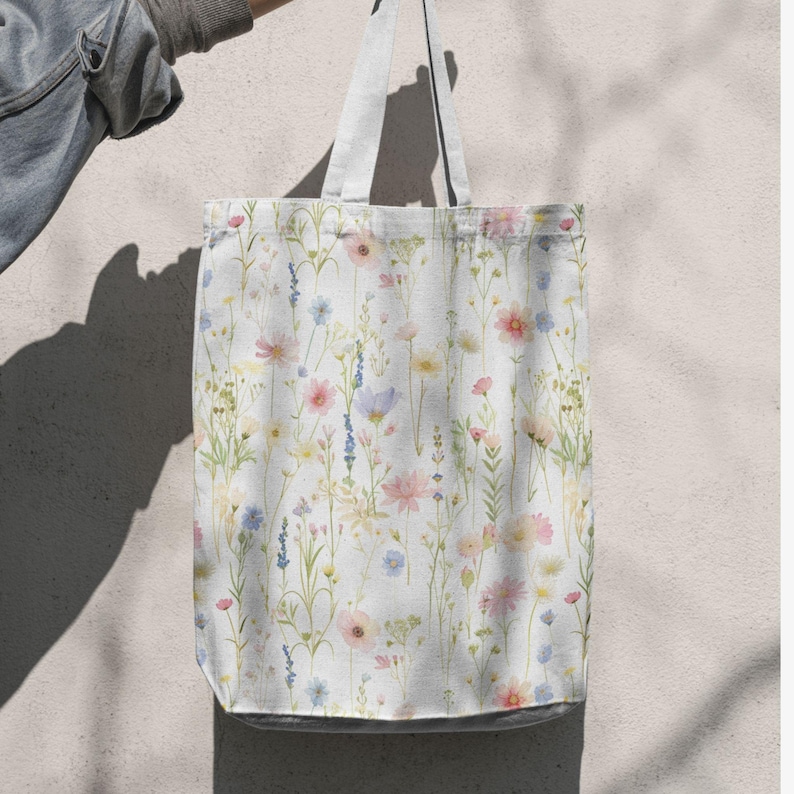 May include: A white tote bag with a floral pattern featuring pink, yellow, blue, and purple wildflowers. The bag has white handles and a light gray bottom. The bag is held up against a light-colored wall.