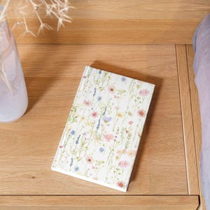 May include: A floral-patterned journal with a white cover featuring a watercolor design of wildflowers in pink, blue, and yellow. The journal is rectangular and rests on a wooden surface, suggesting a desk or bedside table.