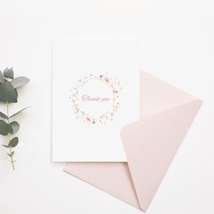 K&ouml;nnte beinhalten: Eine wei&szlig;e Gru&szlig;karte mit dem mittig aufgedruckten Text Thank you, umgeben von einem hexagonalen Kranz aus zarten rosa und blauen Aquarellblumen, begleitet von einem passenden hellrosa Umschlag und einem Zweig aus gr&uuml;nem Eukalyptus.