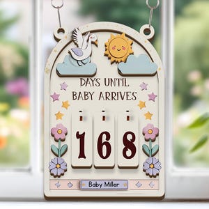 May include: Wooden baby countdown sign with a swan, sun, and cloud design. The sign reads "DAYS UNTIL BABY ARRIVES" and displays "168" days. The bottom of the sign says "Baby Miller". The sign is decorated with stars and flowers.