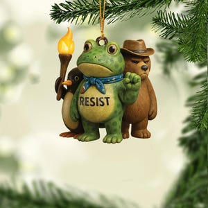 May include: A whimsical Christmas ornament featuring a green frog with the word "RESIST" on its belly, a penguin holding a torch, and a bear wearing a hat. The frog has a blue scarf. The ornament is suspended by a gold string.
