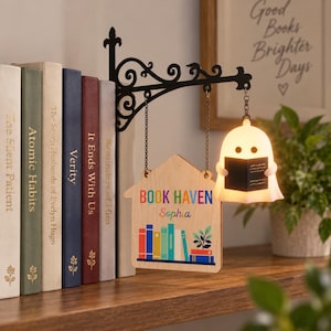 May include: A black metal wall bracket holds a wooden house-shaped sign and a glowing ghost-shaped lamp. The sign features the text BOOK HAVEN and the name Sophia, with a colorful illustration of books and a plant. The ghost lamp holds a small book.