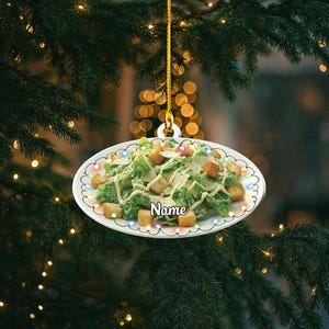 May include: A white, oval-shaped Christmas ornament featuring a Caesar salad design. The salad includes green lettuce, croutons, and a creamy dressing. The word "Name" is printed on the ornament, which is trimmed with colorful lights.