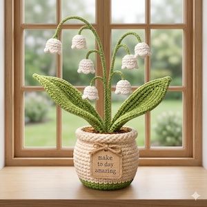May include: A crocheted lily of the valley plant in a beige pot with green trim. The plant features white bell-shaped flowers, green leaves, and stems. A tag on the pot reads "make to day amazing."