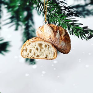 May include: A bread-shaped Christmas ornament, featuring a loaf and a slice, with the word "Name" on the loaf. The ornament is suspended by a gold string against a backdrop of evergreen branches and a snowy background.
