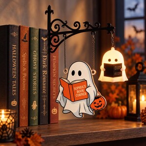 May include: A black metal wall bracket holds a hanging wooden ghost illustration reading an orange book labeled Sophia's Book Corner and a small glowing white ghost lamp. The display is positioned next to a row of five hardcover books.