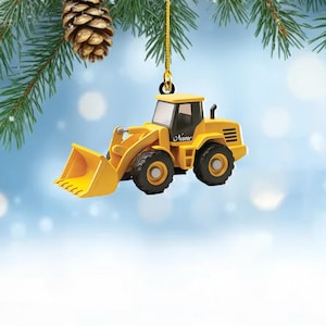 May include: Yellow construction loader Christmas ornament with black tires and a bucket. The ornament has the word "Name" on the side. The ornament is hanging from a gold string, with a pine cone and pine needles in the background.