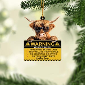 May include: A brown Highland cow ornament wearing sunglasses, holding a yellow and black sign with the text "WARNING VEHICLE RULES". The ornament is suspended by a gold string, perfect for holiday decor.
