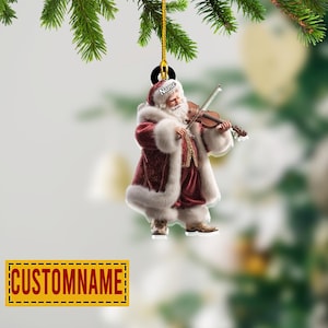 May include: A Christmas ornament featuring Santa Claus playing a violin. Santa is dressed in a red and white fur-trimmed suit. The ornament has a gold string for hanging and a yellow rectangle with the text "CUSTOMNAME".