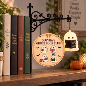 May include: A black metal wall-mounted bracket holds a round wooden sign that reads SOPHIA'S GHOST BOOK CLUB with four ghost illustrations reading books. A small, illuminated white ghost figure holding a book hangs from the bracket by a chain.