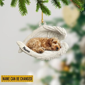 May include: A personalized Christmas ornament featuring a sleeping, light brown Cockapoo dog nestled in white angel wings. The ornament has a gold string for hanging and the text "Name" is written on the wing. The text "NAME CAN BE CHANGED" is at the bottom.
