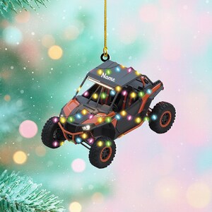 May include: An orange and black UTV ornament with a string of colorful lights. The ornament has the word "Name" on the roof. The UTV has black wheels and is hanging from a gold string.