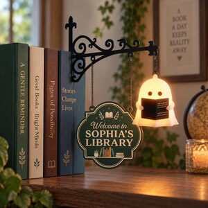 May include: A black metal wall-mount bracket holds a hanging sign that reads Welcome to SOPHIA'S LIBRARY with a book graphic. Beside it, a small white ghost-shaped lamp glows, holding a black book. The sign hangs by chains from the bracket.