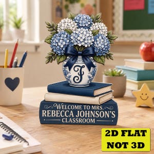 May include: A flat, 2D decorative sign featuring a stack of three books with a blue and white vase of blue and white hydrangeas on top. The bottom book is dark blue with the text "WELCOME TO MRS. REBECCA JOHNSON'S CLASSROOM" and a navy bow.