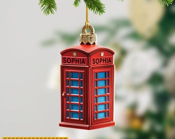 May include: A red Christmas ornament shaped like a British telephone box, with blue window panels. The name "SOPHIA" is printed on the top. The ornament hangs from a gold-colored loop and a gold string. The text "NAME CAN BE CHANGED" is at the bottom.
