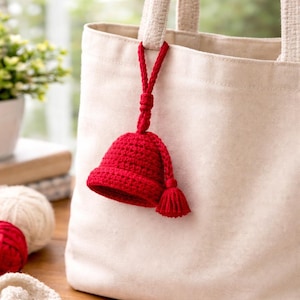 May include: A handmade red crochet bell charm with a tassel, hanging from the handle of a canvas tote bag. The bell is attached to a braided cord. The background includes yarn balls and a plant in a white pot.