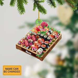 May include: A wooden sushi platter Christmas ornament. The ornament features a variety of sushi rolls, nigiri, and garnishes, with a string of lights. The ornament has a gold string for hanging and the word "Name" is printed on the platter.