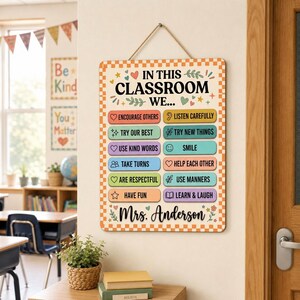 May include: A rectangular classroom wall sign with a checkered orange and white border. The text reads IN THIS CLASSROOM WE... followed by a list of rules like ENCOURAGE OTHERS, LISTEN CAREFULLY, and TRY OUR BEST. The sign is personalized with Mrs. Anderson.