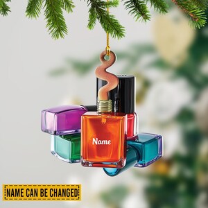 May include: A Christmas ornament shaped like a collection of nail polish bottles in various colors, including orange, purple, green, and teal. The orange bottle has the word "Name" printed on it. The ornament is hanging from a green pine branch.