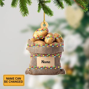 May include: A burlap sack ornament filled with potatoes, adorned with colorful string lights. The ornament has a small tag that says "Name" and a separate tag that says "Name can be changed". The ornament is hanging from a green branch.