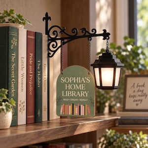 May include: A black metal wall-mounted bracket with a hanging lantern and a green sign that reads "SOPHIA'S HOME LIBRARY" and "READ • DREAM • REPEAT" above a graphic of colorful books, positioned next to a row of hardcover books on a shelf.