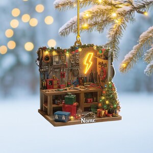 May include: A Christmas ornament shaped like a workshop, featuring tools, a small Christmas tree, and a neon lightning bolt. The ornament has a wooden workbench, a blue toolbox, and several wrapped gifts. The word "Name" is printed on the ornament.