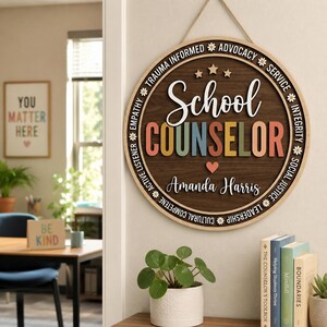 May include: A circular wooden wall sign featuring the text School Counselor in white and multicolored block letters. The name Amanda Harris is printed below. The border includes the words Trauma Informed, Advocacy, Service, Integrity, Social Justice, Leadership, Cultural Competence, Active Listener, and Empathy separated by small white daisy icons.