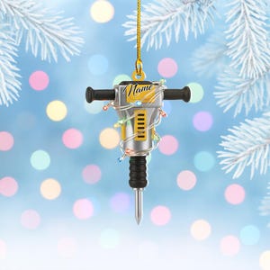 May include: A silver and black jackhammer Christmas ornament with the word "Name" in gold script. The ornament is wrapped with a string of colorful lights and hangs from a gold string. The background is a blurred blue with white and yellow bokeh.
