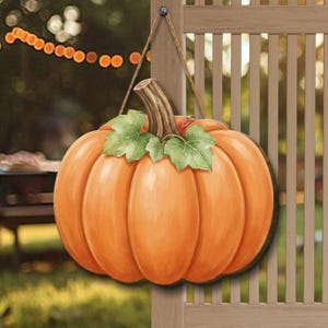 May include: A decorative hanging sign shaped like a round orange pumpkin with a brown stem and green leaves. The pumpkin features a smooth, painted surface with visible segments and is suspended by a thin, natural-colored twine cord.