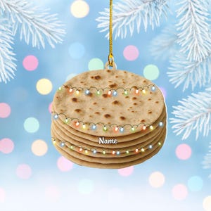 May include: A stack of flatbread-shaped Christmas ornament with a string of colorful lights. The ornament has a gold string for hanging and the word "Name" is printed on the front. The background is a blurred blue with white frosted branches.
