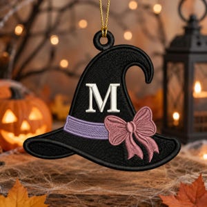 May include: A black embroidered witch hat ornament featuring a white letter M in the center, a light purple band, and a dusty rose-colored bow. The ornament hangs from a gold string and has a curved, pointed top with a wide brim.