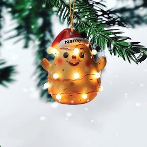 May include: A festive Christmas ornament shaped like a smiling character, wearing a Santa hat with the word "Name" on it. The ornament is wrapped in a string of warm white lights and hangs from a gold string against a backdrop of evergreen branches.