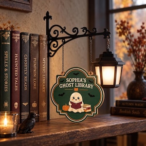 May include: A decorative hanging sign featuring the text 'SOPHIA'S GHOST LIBRARY' with a cartoon ghost reading a book, a pumpkin, and bats. The sign is suspended by chains from a black metal bracket that also holds a small, illuminated lantern.
