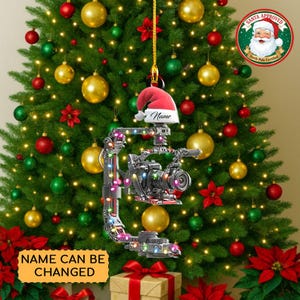 May include: A Christmas tree ornament featuring a camera design with a Santa hat. The ornament has a customizable name and is decorated with colorful lights. The tree is adorned with red, green, and gold ornaments and poinsettias.