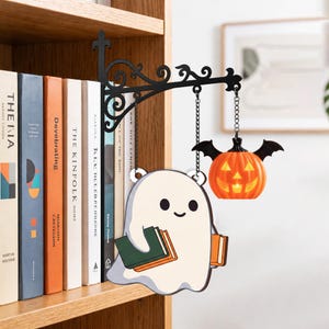 May include: A black metal wall-mounted bracket with scrollwork, featuring two hanging chains. One chain holds a flat, white ghost illustration holding books, and the other chain holds a small, orange, bat-winged jack-o'-lantern light.