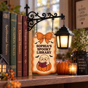 May include: A hanging wooden sign with a black metal bracket and lantern. The sign features an orange bow, the text "SOPHIA'S SPOOKY LIBRARY," and a cartoon ghost wearing glasses while reading a book inside a pumpkin, surrounded by small bats.