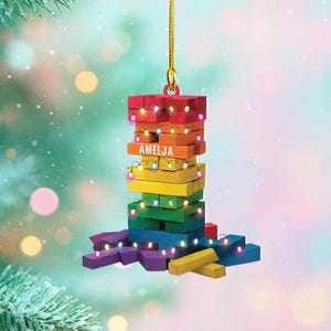May include: A colorful, wooden Christmas ornament shaped like a tower, with the name "AMELJA" on it. The ornament is made of stacked blocks painted in rainbow colors and adorned with tiny lights. It hangs from a gold string.