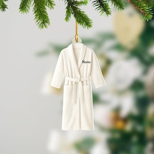 May include: A cream-colored robe ornament with the word "Name" embroidered on the lapel. The robe has a belt and pockets, and is hanging from a gold string. The ornament is suspended in front of a blurred background of greenery.