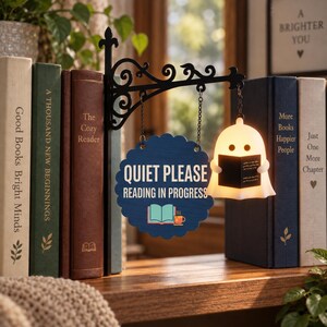 May include: A black metal wall-mount bracket holds a blue scalloped sign that reads QUIET PLEASE READING IN PROGRESS with a book and mug icon. Next to it, a small white ghost-shaped lamp holds a black book, illuminated from within.