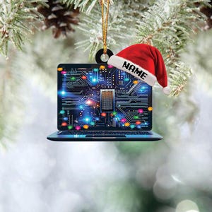May include: A laptop-shaped Christmas ornament with a circuit board design on the screen. A red Santa hat with the word "NAME" is on top. The ornament hangs from a gold string against a blurred background of pine needles.
