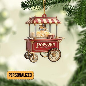 May include: A red and gold popcorn cart Christmas ornament with a striped awning. The cart has the word "Popcorn" on the front and is filled with popcorn. The ornament is personalized with the word "Name".