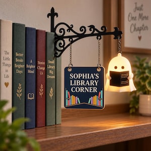 May include: A black metal wall bracket holds a square sign reading SOPHIA'S LIBRARY CORNER with a colorful book graphic and a small, illuminated white ghost figure holding a book, all suspended by chains next to a row of five hardcover books.
