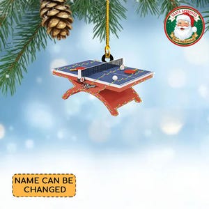 May include: A Christmas ornament shaped like a ping pong table, with a red base and blue playing surface. The ornament includes a net, paddles, and ping pong balls. The text "Name" is written on the table. The ornament is hanging from a gold string.