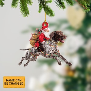 May include: A Christmas ornament featuring Santa Claus riding a German Shorthaired Pointer dog. The dog is brown and white with a festive garland of lights. Santa wears a red suit and hat, carrying a sack of gifts. The ornament has a gold string for hanging.
